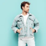 portrait-handsome-confident-stylish-hipster-lambersexual-model-with-curly-hairstyle-sexy-man-dressed-jeans-jacket-fashion-male-isolated-blue-wall-studio